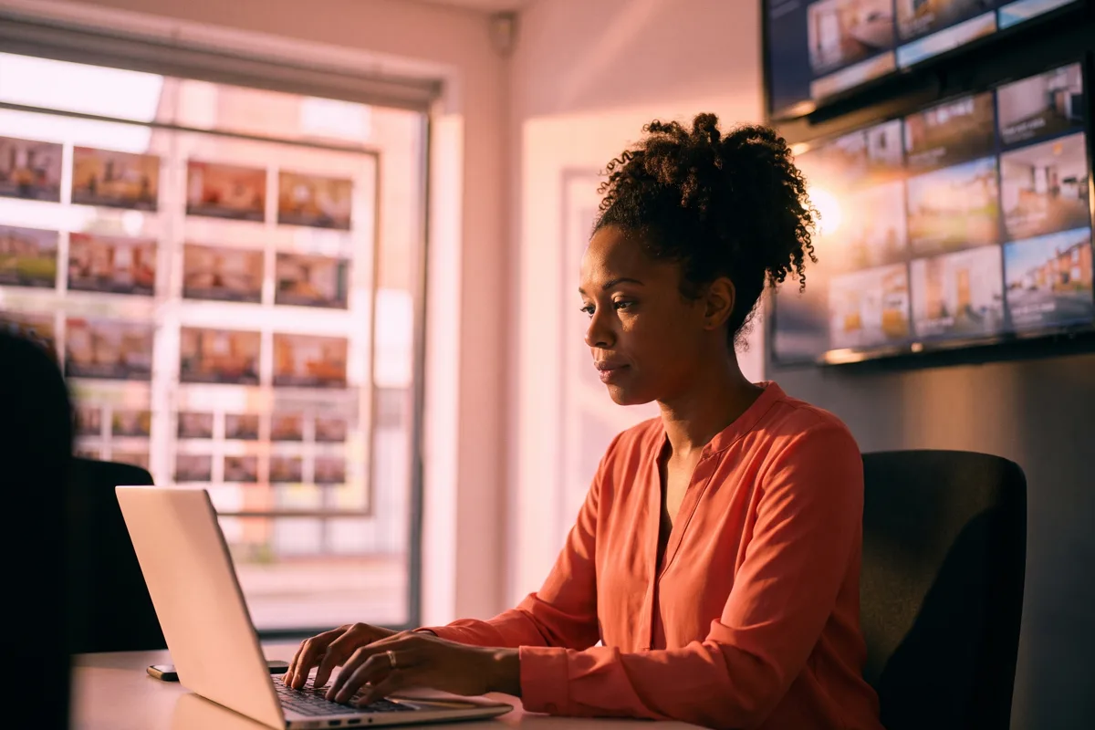 A marketing manager works at a laptop in a modern multi-branch estate agency office, two large screens glowing behind her with rows of article thumbnails