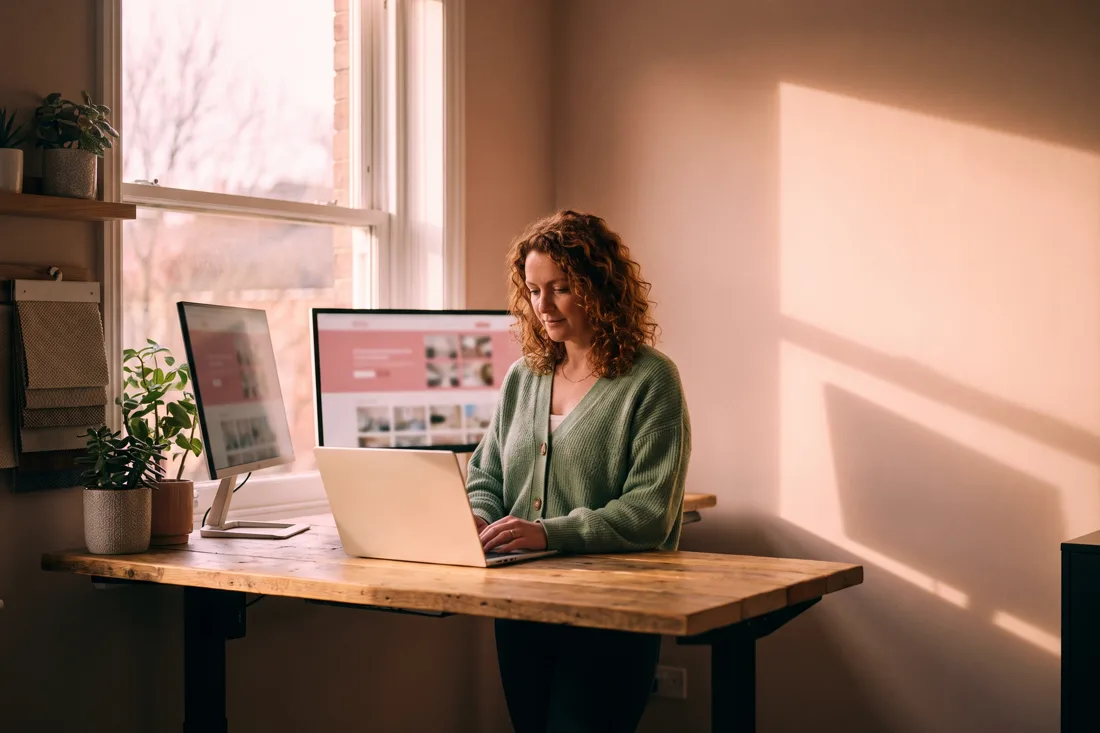 An estate agent at a standing desk reviewing their agency website's listings page on a laptop and monitor
