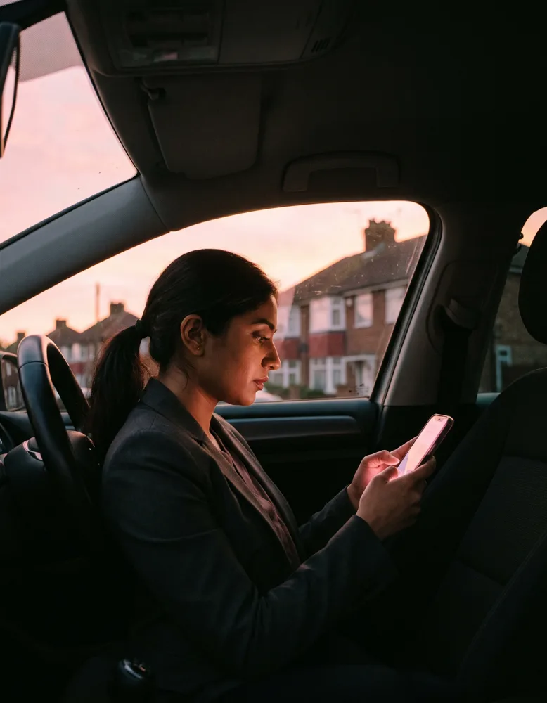 An estate agent sitting in a parked car outside a British semi-detached house at dusk, phone glowing as they send a document