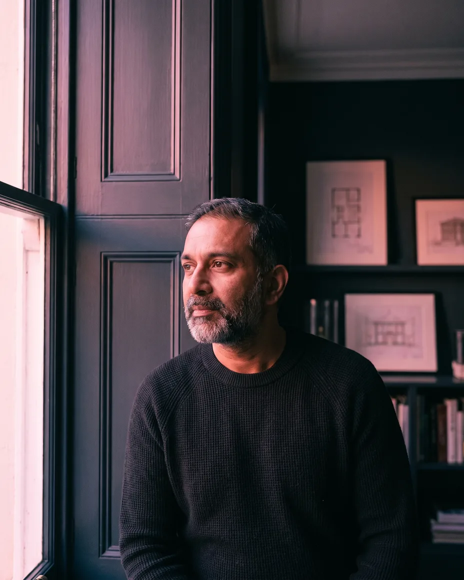 A thoughtful portrait of a surveying firm principal by a window in soft pink light, dark panelling behind him