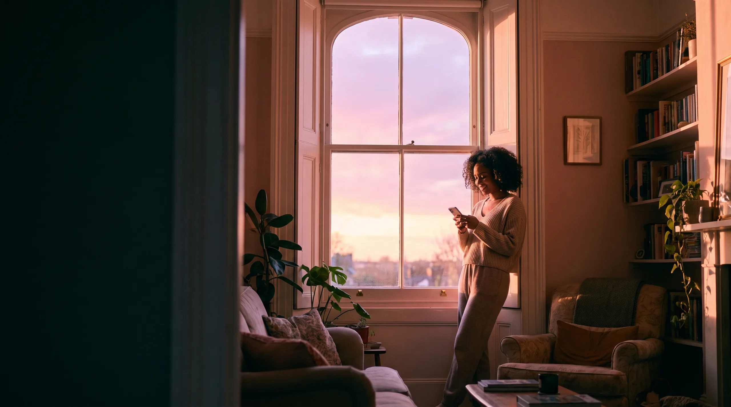 A homeowner stands beside a tall sash window in soft pink morning light, checking her survey on her phone
