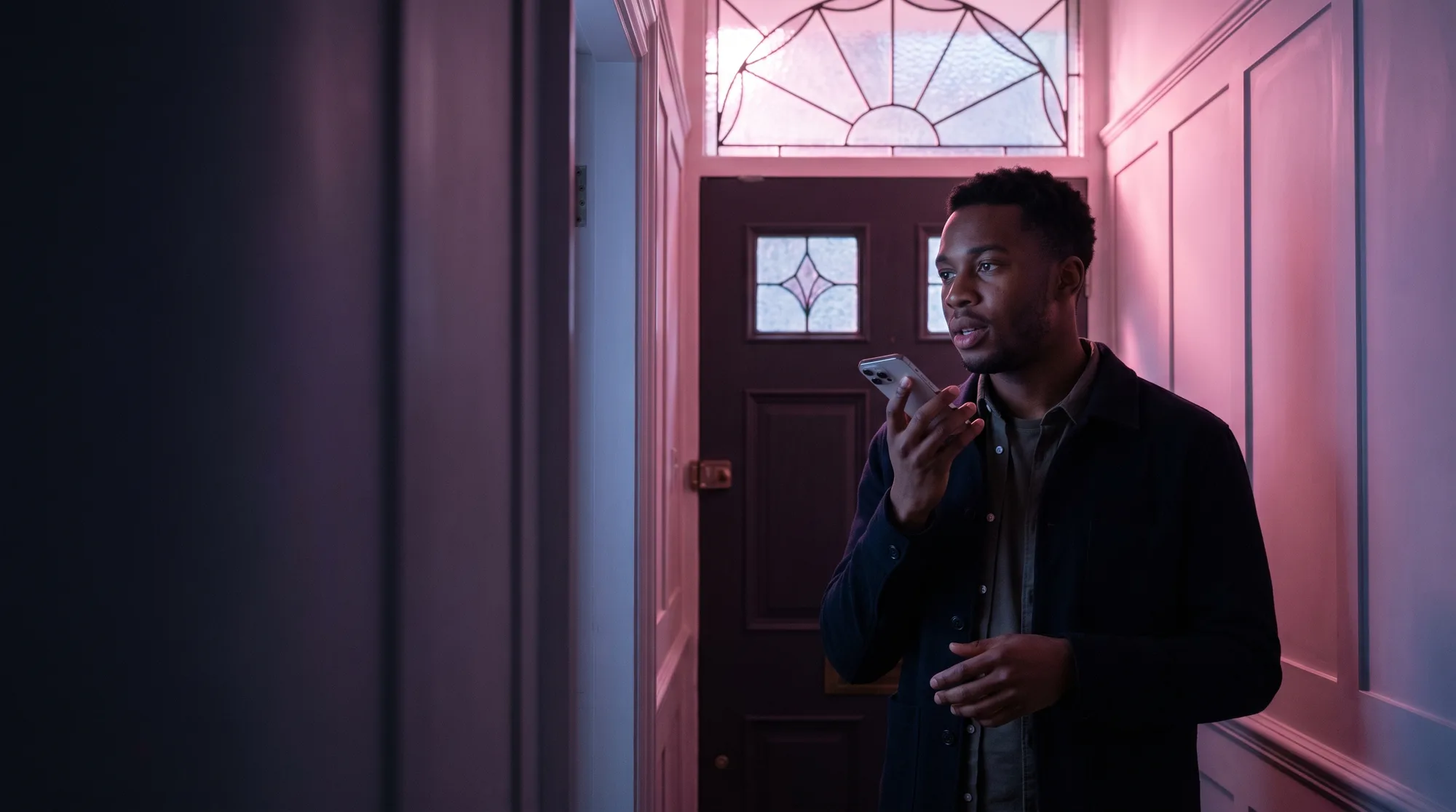 A surveyor stands in the hallway of a period house at dusk, asking his phone a question, pink light through the fanlight behind him