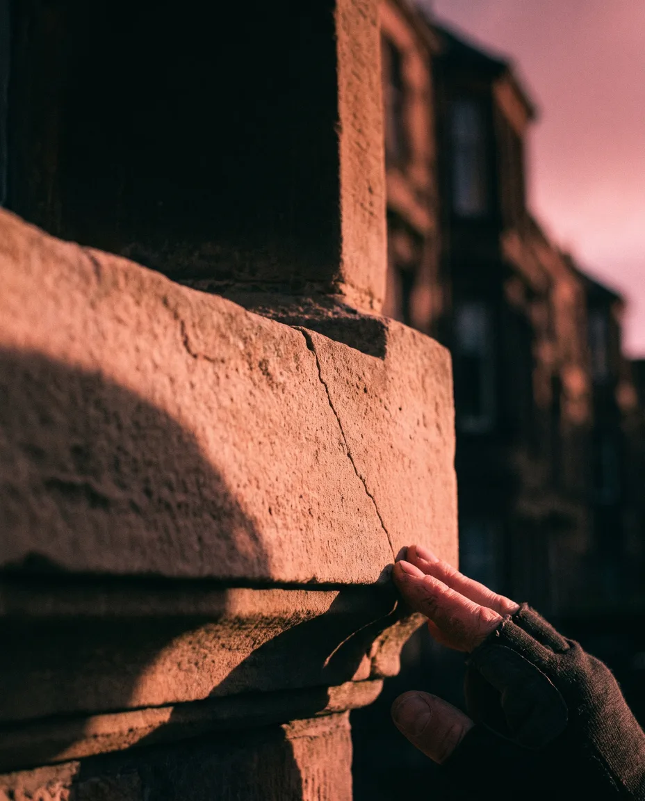 A surveyor's hand tracing a fine hairline crack across a weathered stone window lintel in warm pink evening light