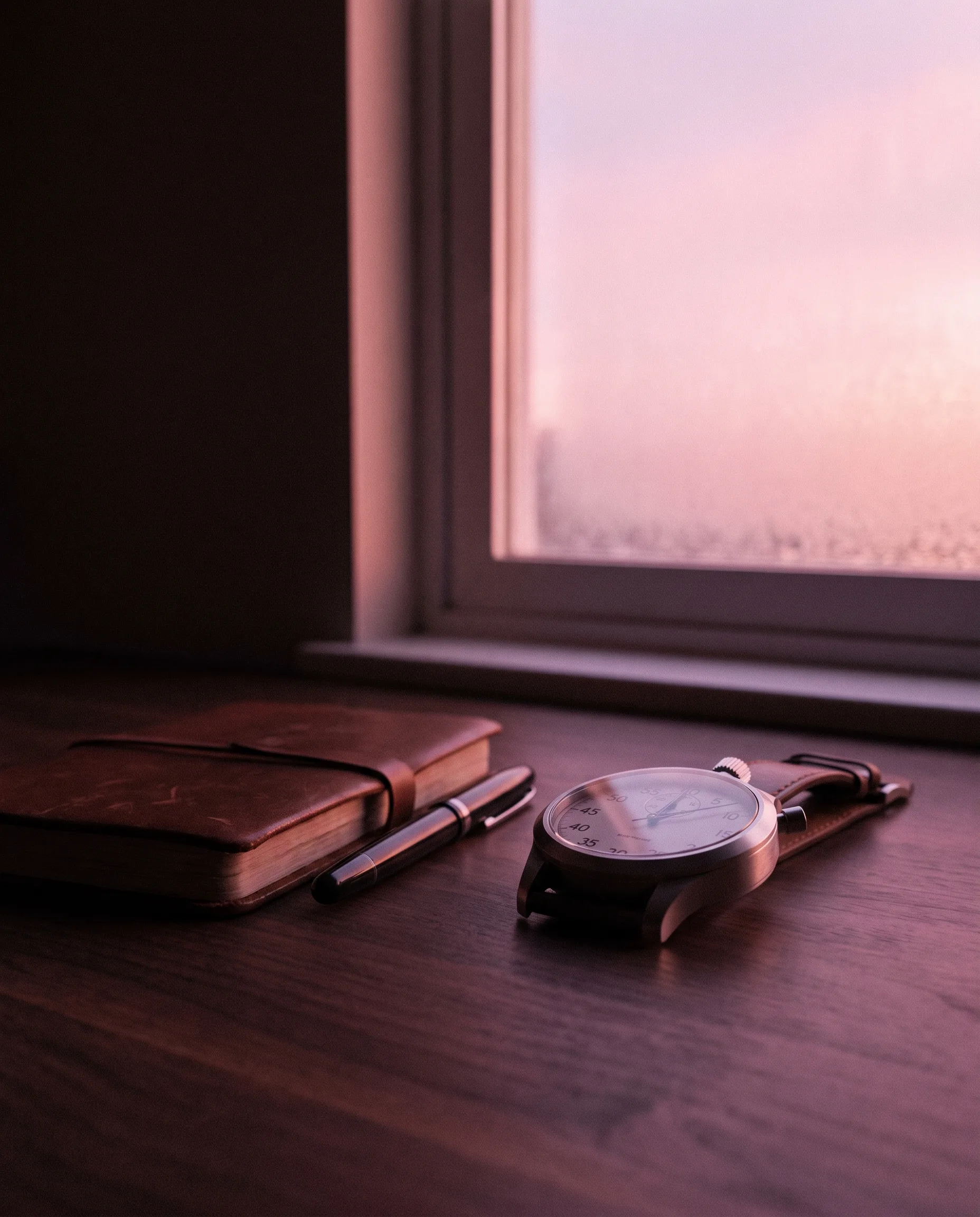 A modern analogue stopwatch on a leather strap beside a notebook and pen on a dark walnut desk, washed in rose-pink dawn light from the window
