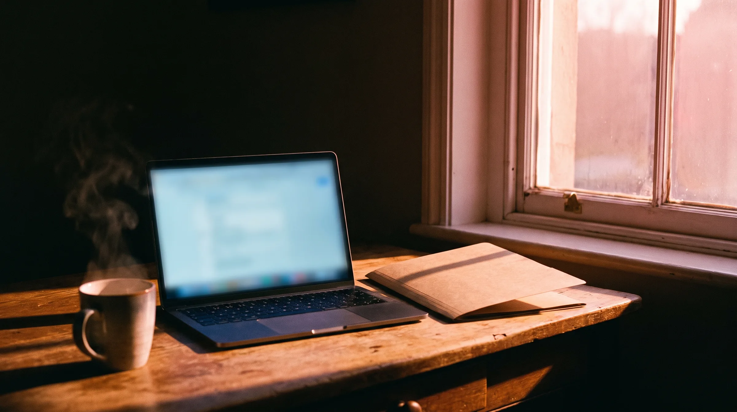 A home-office desk at dawn — an open property folder beside a glowing laptop, pink sunrise light through the sash window
