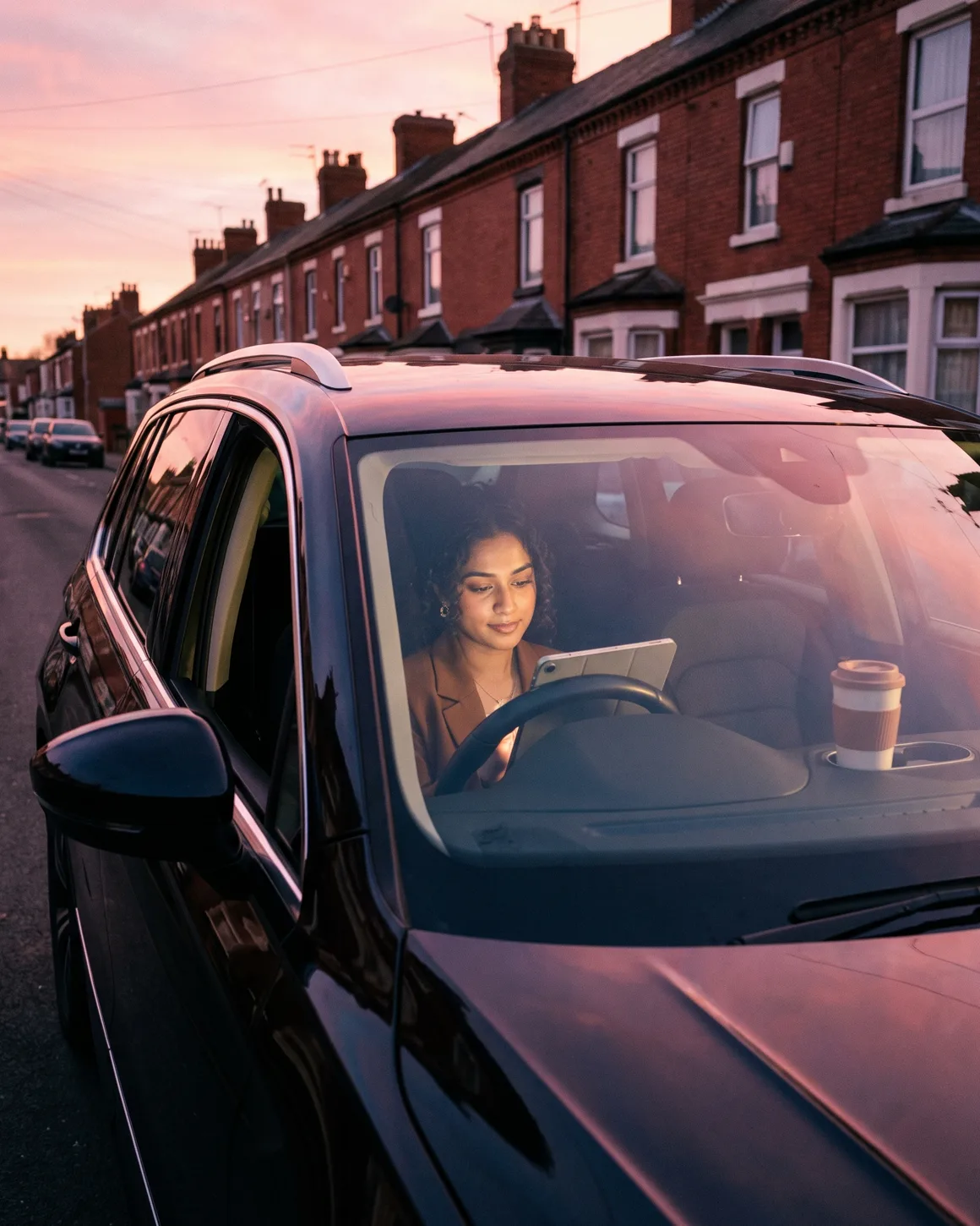 A surveyor in her parked car at sunrise outside a red-brick Victorian terrace, reviewing the property dossier on a tablet, coffee in the cupholder