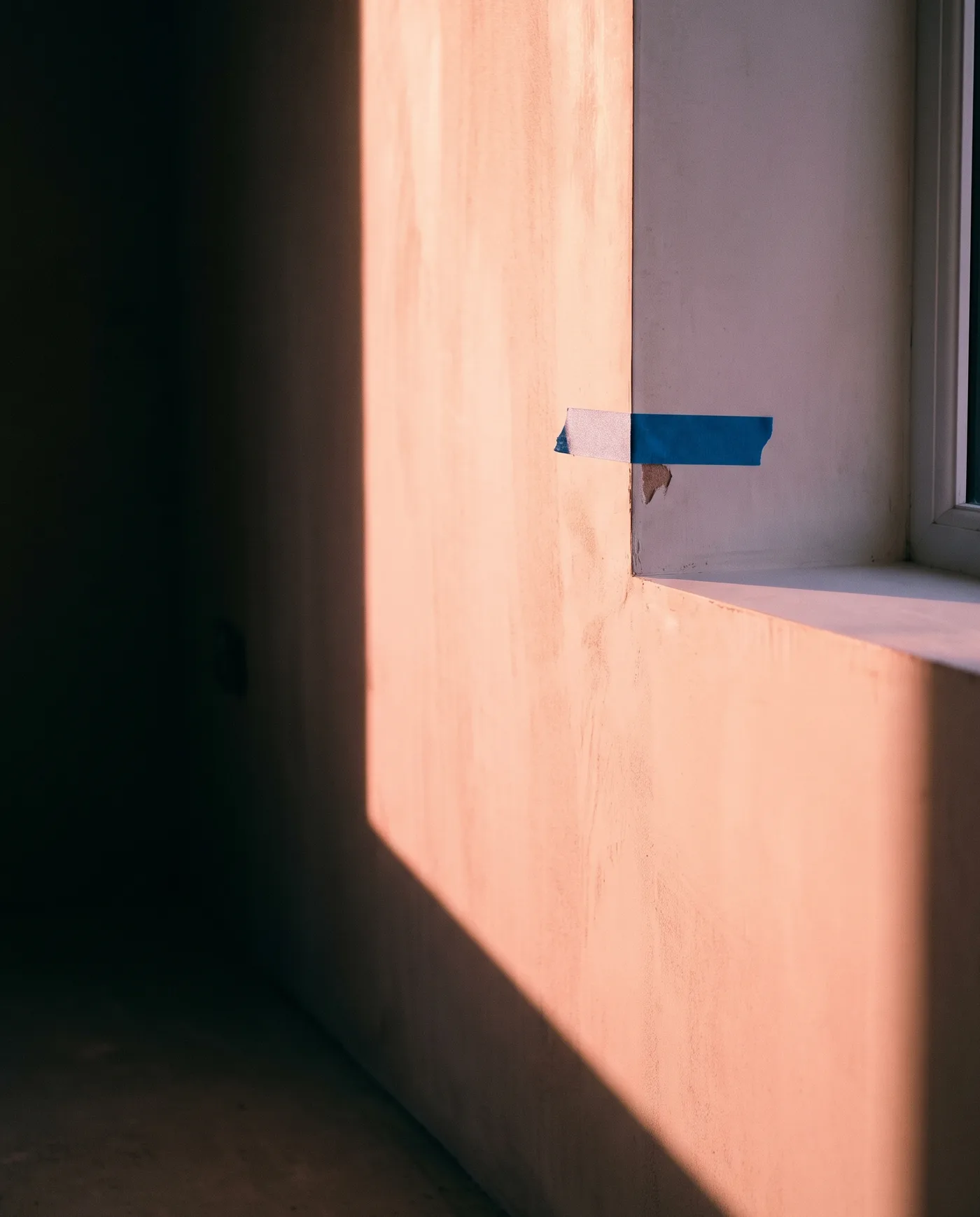 A strip of tape marking a chipped-paint defect on the corner of a window reveal in a freshly plastered new-build room, pink dawn light raking across the wall