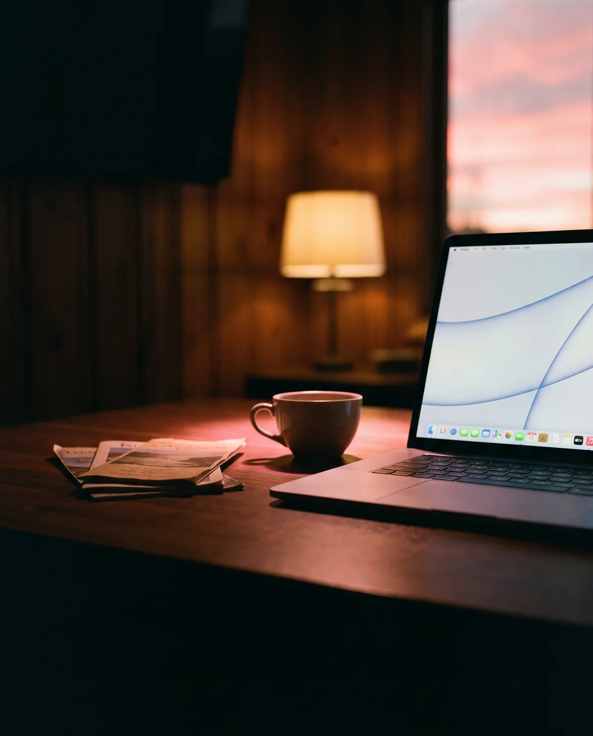 A laptop glowing on a dark desk at dusk, a stack of old printed brochures pushed aside into the shadow beside a cup of coffee