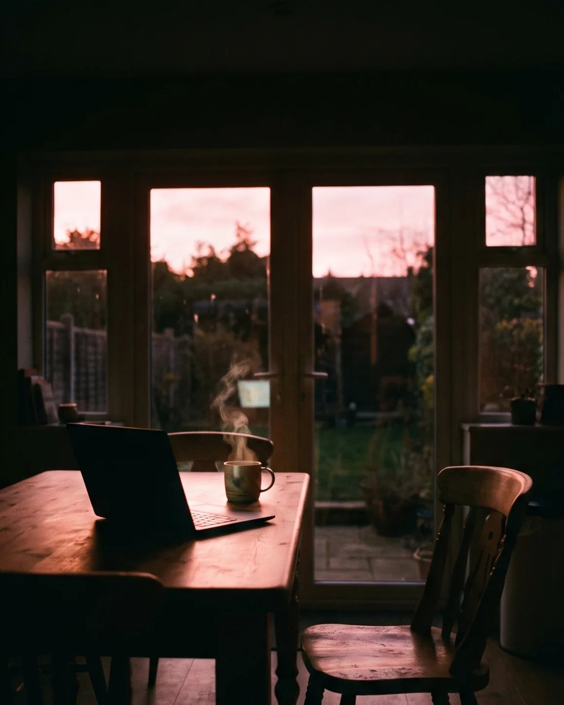 A kitchen table at dusk with a laptop open and a mug of tea steaming, pink evening light in the garden beyond the doors